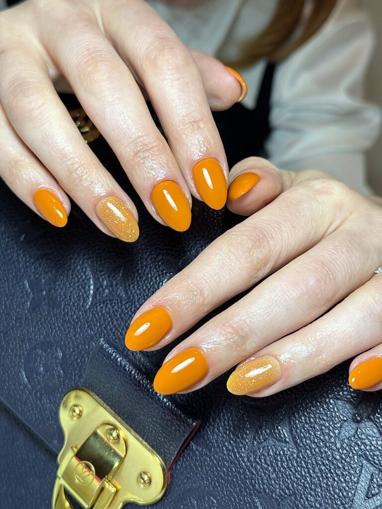 Glossy orange almond-shaped manicure with a gold glitter accent
